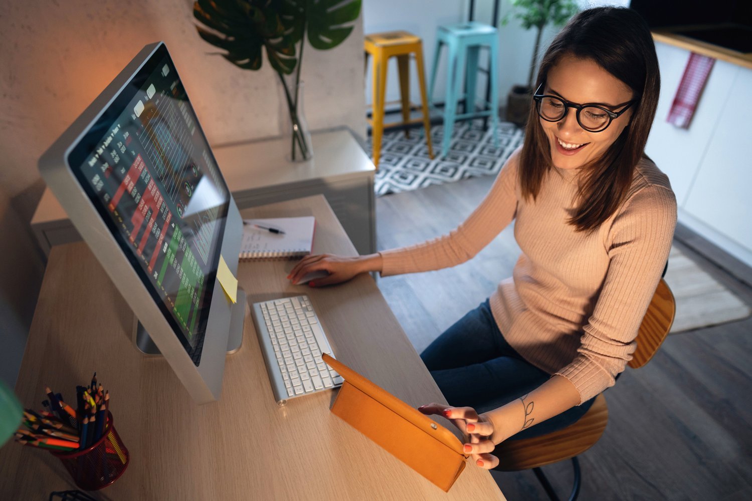 High-angle view of a young woman trading cryptocurrency on a computer and digital tablet High-angle view of a young Caucasian woman tracking and trading stocks, using a computer and digital tablet at home. Stock exchange, investment, and financial trading.