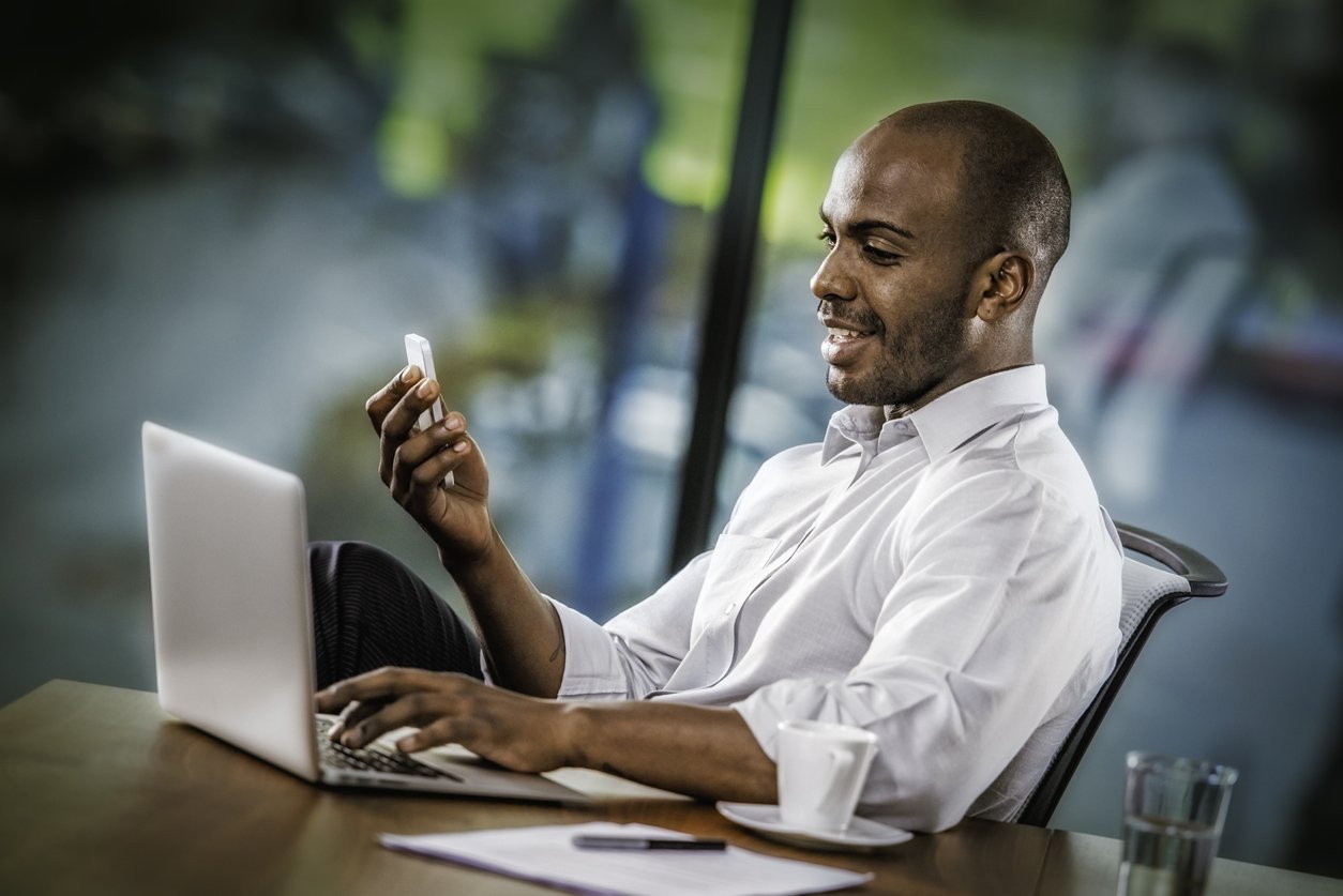 Smiling Businessman Having a Video Call Young African American businessman having a video call on a mobile phone while using laptop in his office, tilt image.